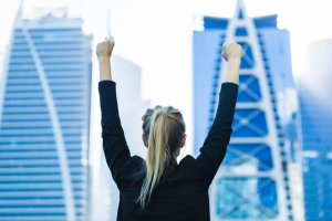 A person with long blonde hair, wearing a black jacket, stands with arms raised in a victory pose against a backdrop of modern skyscrapers in a cityscape. As they bask in the clear sky and the blue and white designs of the buildings, their mind is already journaling this triumphant moment.