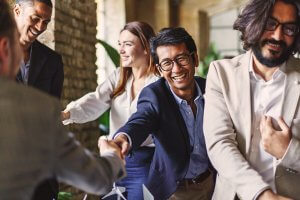 A diverse group of four people, dressed in business casual attire, are smiling and shaking hands in a bright office setting. They appear to be celebrating or concluding a successful, insightful meeting, with a model wind turbine visible on the table in front of them.