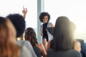 A woman with curly hair, wearing a sleeveless vest and holding a tablet, smiles while showcasing her leadership skills to an attentive audience. She stands in front of a bright window. One audience member has their hand raised, while others are seated, focusing on her.