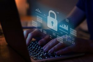 A person is typing on a laptop with a holographic image of a padlock and data icons, representing cybersecurity while remote working. The scene is dimly lit, emphasizing the glow of the laptop screen and the security icons.