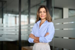 A confident individual, embodying strong values, stands in an office setting with long hair, wearing a light blue shirt and light pants. Their arms are crossed as they smile softly. The background features glass walls with frosted stripes.