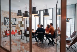 A diverse group of people sits around a conference table in a modern office with large glass walls, embodying the company's values. They engage in a meeting, focused on a screen on the wall. Hanging lights and contemporary decor create a professional atmosphere.
