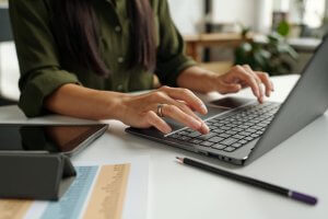 A person in a green top types on a laptop at a desk. Nearby, there's a tablet, a printed chart with colorful sections, and a pencil. The setting, reminiscent of a bright, modern government office, buzzes with productivity.