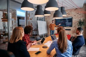 A group of five people are in a modern office, gathered around a table with laptops and papers. A man stands near a whiteboard and a large screen displaying code, gesturing as he presents innovative service solutions. Hanging lamps and a brick wall complete the setting.