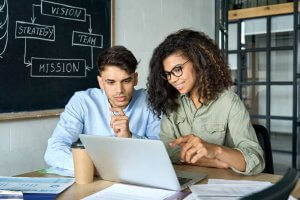 A man and a woman sit at a desk looking at a laptop. The woman, smiling, points at the screen as they discuss key DEI initiatives. They are surrounded by papers and a coffee cup. A chalkboard behind them displays the words: "Vision," "Strategy," "Team," and "Mission.