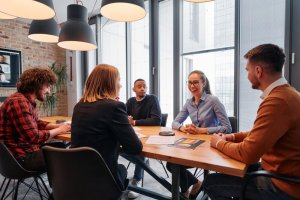 A diverse group of five people are engaged in a meeting around a wooden table in a modern office, demonstrating leadership as they discuss business documents. A laptop is open, while large windows and hanging lights create a bright atmosphere.