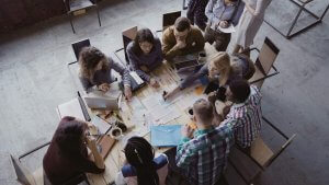 A group of diverse people sit around a large table, embodying DEI principles as they engage in discussion and work. Papers, laptops, and stationery are spread across the table. The setting appears to be a collaborative office or classroom environment.