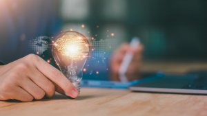 A person holds a glowing light bulb with a digital brain pattern inside, symbolizing innovation and ideas interwoven with modern culture. The scene features a blurred background with a tablet and stylus on a wooden desk, capturing the essence of creative evolution.