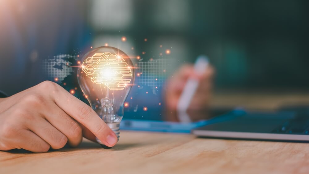 A person holds an illuminated light bulb with a brain design inside, symbolizing an idea fueled by leadership. The background features a blurred view of a tablet and laptop on a wooden table, suggesting a focus on technology and innovation. A person holds an illuminated light bulb with a brain design inside, symbolizing an idea fueled by leadership. The background features a blurred view of a tablet and laptop on a wooden table, suggesting a focus on technology and innovation.