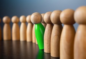 A row of wooden figures stands upright, with one bright green figure leaning slightly, contrasting with the others. The background is a soft gradient, much like the emergence of AI emphasizing the uniqueness of the vibrant green figure among its wooden counterparts.