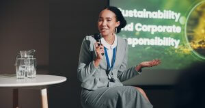 A leader in a gray suit speaks into a microphone while seated on a stool. Behind them, a presentation slide reads "Sustainability and Corporate Responsibility." A glass pitcher of water rests on a small round table beside them.