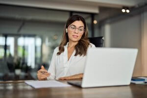 A woman in a white blouse sits at a desk, exemplifying leadership as she engages in a video call. She's holding a pen with papers nearby, her focused demeanor reflecting her role. The modern office, with its sleek glass partitions, underscores the professional atmosphere.