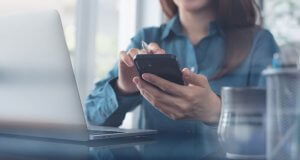 A person in a blue shirt sits at a desk, holding a smartphone in one hand and typing with the other. An open laptop is in front of them, and a pen holder is visible in the background. The scene suggests a professional work environment, possibly within a government office.