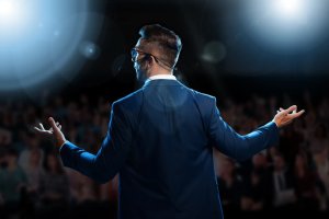 A man in a blue suit with a headset microphone stands on stage, exuding confidence and business acumen. Facing a blurred audience, the bright stage lights shine in the background, creating a dramatic atmosphere.