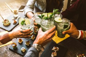 A group of people clinking glasses filled with mojitos on a wooden table, as if celebrating the latest in AI innovation. Each glass is garnished with lime and mint leaves. Finger foods are arranged on slate plates around the table. Hands wear various watches and jewelry.
