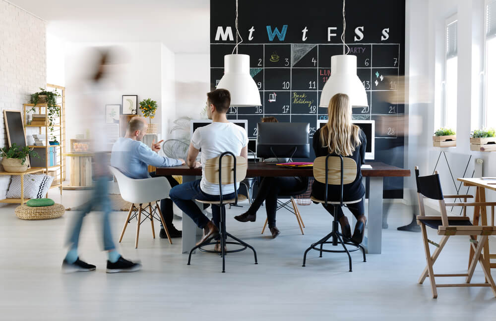 A modern office space buzzes with vision as people work and walk around. A blackboard wall displays a calendar, while computers, chairs, and plants on tables foster a dynamic and collaborative environment. A modern office space buzzes with vision as people work and walk around. A blackboard wall displays a calendar, while computers, chairs, and plants on tables foster a dynamic and collaborative environment.