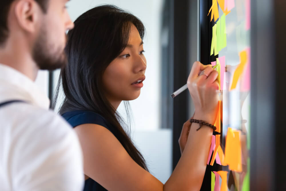 A woman is writing on colorful sticky notes placed on a blackboard, while a man stands beside her in a modern office, embodying the collaborative spirit of the public sector as they focus on organizing or planning ideas. A woman is writing on colorful sticky notes placed on a blackboard, while a man stands beside her in a modern office, embodying the collaborative spirit of the public sector as they focus on organizing or planning ideas.