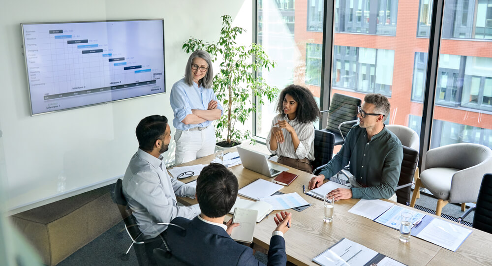 A group of five colleagues in a modern office discusses a hybrid project. A woman stands near a screen displaying a timeline chart, while others sit around a table with laptops, papers, and glasses of water. Large windows reveal the cityscape in the background. A group of five colleagues in a modern office discusses a hybrid project. A woman stands near a screen displaying a timeline chart, while others sit around a table with laptops, papers, and glasses of water. Large windows reveal the cityscape in the background.