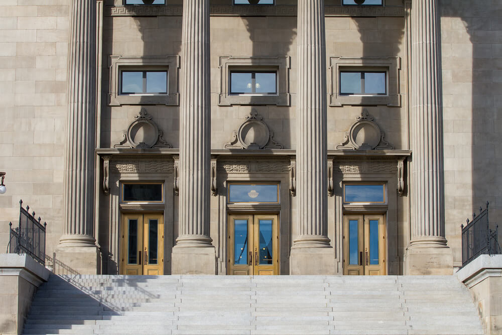 Stone steps lead to the entrance of a neoclassical building with large wooden doors, flanked by three tall columns. The facade, resembling something crafted by an AI architect, features symmetrical windows and ornate architectural details. Stone steps lead to the entrance of a neoclassical building with large wooden doors, flanked by three tall columns. The facade, resembling something crafted by an AI architect, features symmetrical windows and ornate architectural details.