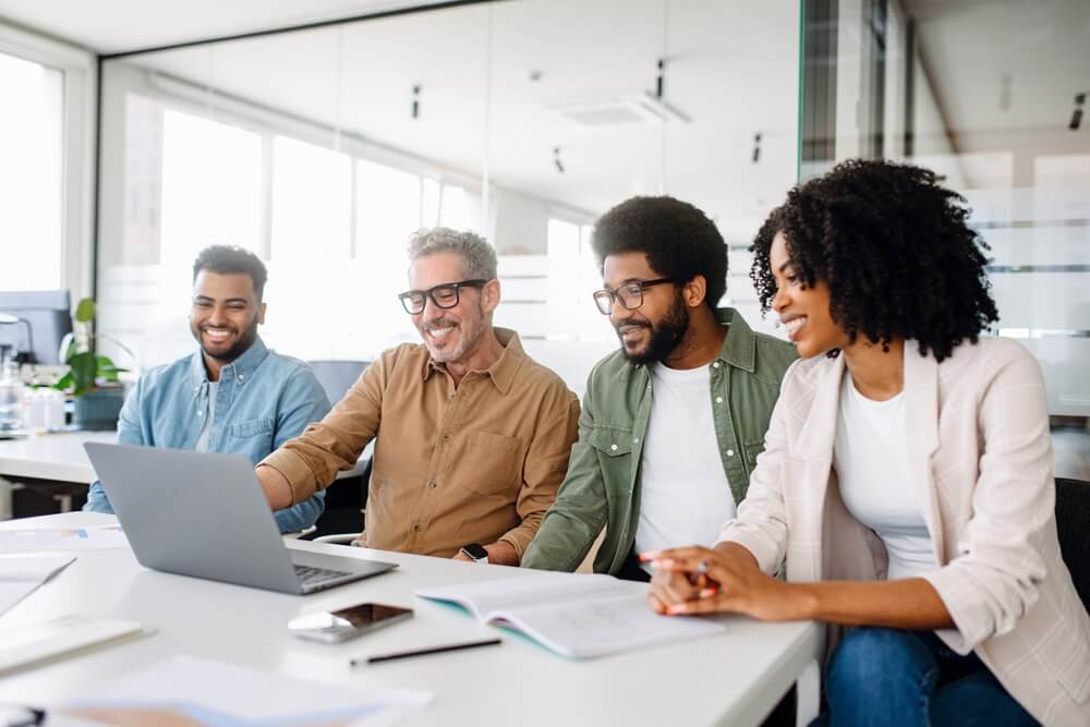 A diverse group of four people sit around a table with a laptop, smiling and engaged in a collaborative work environment. The modern office, with its large windows, sets the scene for leadership to flourish as books and documents spread across the table inspire innovative ideas. A diverse group of four people sit around a table with a laptop, smiling and engaged in a collaborative work environment. The modern office, with its large windows, sets the scene for leadership to flourish as books and documents spread across the table inspire innovative ideas.