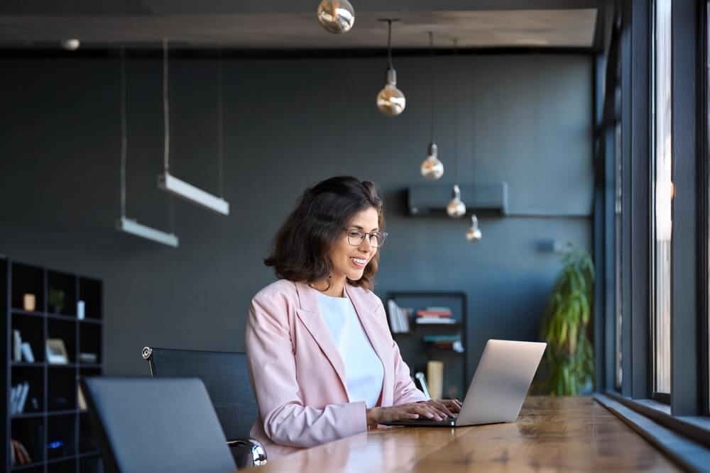 A woman in a pink blazer sits at a long wooden table, working on her laptop in a bright, modern workplace. She appears focused and happy amidst large windows, pendant lights, and a well-stocked bookshelf in the background. A woman in a pink blazer sits at a long wooden table, working on her laptop in a bright, modern workplace. She appears focused and happy amidst large windows, pendant lights, and a well-stocked bookshelf in the background.