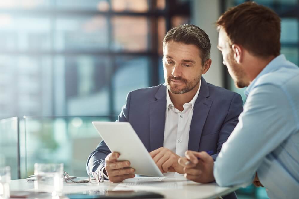 Two men sitting at a table in a modern office workplace are engaged in a discussion. One is holding a tablet, and they both appear focused on its screen. Sunlight streams through large windows in the background, creating a bright, professional atmosphere. Two men sitting at a table in a modern office workplace are engaged in a discussion. One is holding a tablet, and they both appear focused on its screen. Sunlight streams through large windows in the background, creating a bright, professional atmosphere.