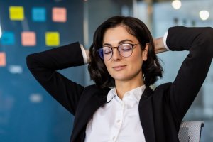 A woman in a business suit, embodying leadership, leans back in her chair with hands behind her head and eyes closed, exuding relaxation. In the background, a glass wall adorned with colorful sticky notes is visible.