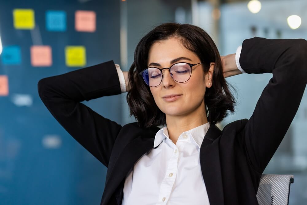 A woman in a business suit, embodying leadership, leans back in her chair with hands behind her head and eyes closed, exuding relaxation. In the background, a glass wall adorned with colorful sticky notes is visible. A woman in a business suit, embodying leadership, leans back in her chair with hands behind her head and eyes closed, exuding relaxation. In the background, a glass wall adorned with colorful sticky notes is visible.