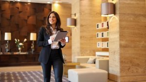 A woman in business attire walks confidently through a modern, stylish lobby, holding a tablet. The lobby features warm lighting, wooden panels, and contemporary decor. She exudes an air of management expertise, appearing focused and professional.