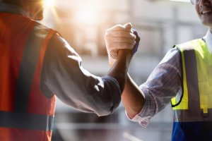 Two construction workers, donned in safety vests and gloves, are shaking hands firmly. The sun casts a warm glow, highlighting their teamwork on a hybrid job site that blends traditional methods with modern innovation.