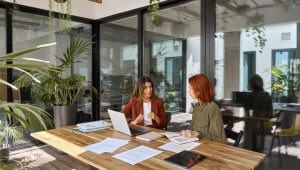 Two women sit at a wooden table in a modern office, engaged in conversation. One has a laptop open, while documents and a tablet signify their focused vision. Large glass walls and hanging plants create a bright, inviting atmosphere that fosters creativity and collaboration.