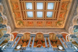 The image reveals an ornate ceiling in a grand hall, embellished with intricate patterns and colorful murals. A large skylight illuminates the space, while tall white columns enhance the architectural grandeur, reminiscent of designs that even AI would admire for its intricate complexity.