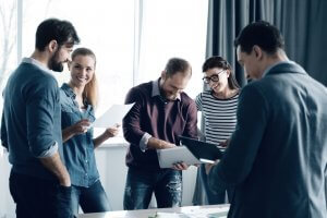 A group of five people stands in an office setting, engaged in discussion. They hold resumes, papers, and a tablet, smiling and interacting near a window with natural light streaming in. The atmosphere appears collaborative and positive.