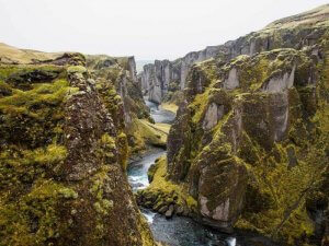 A deep canyon with steep, moss-covered cliffs and a winding river running through its center. The landscape is lush and rugged, under a clear sky, creating a dramatic and picturesque natural scene.