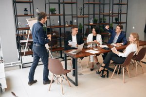 A group of five people gathers in a modern office setting. One stands beside a flipchart, presenting insights akin to crafting the perfect resume. The others sit around a wooden table with laptops and papers, attentively engaged. Shelves adorned with plants provide a serene backdrop.