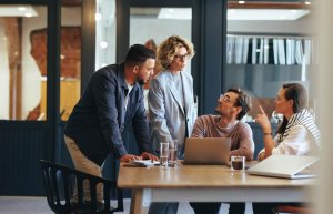 Four people are having a collaborative meeting in an office setting. Three people are seated at a table with laptops and documents, deeply immersed in their work, while one person stands, leaning over the table. They appear engaged in discussion.
