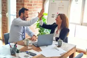 A man and a woman smiling and giving a high-five in a modern office. They sit at a wooden desk with a laptop, papers, cups, and plants around. The room has a brick wall and large windows, allowing natural light to fill the space.