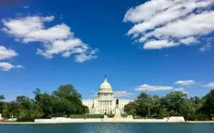 The U.S. Capitol building with its iconic dome is centered under a vibrant blue sky with scattered clouds, surrounded by greenery and situated behind a reflecting pool in the foreground.