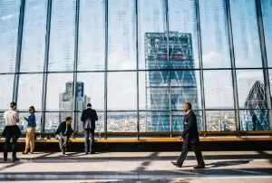 People in business attire are inside a modern building with large glass windows, overlooking a cityscape. Skyscrapers are visible in the background, and a few individuals are standing or walking in the spacious interior.