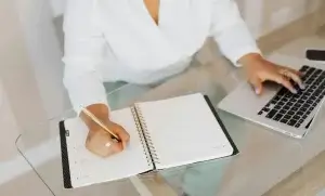 A person wearing a white shirt is sitting at a glass table, writing in an open planner with a gold pen. Their other hand is on a laptop keyboard. The planner is blank, and they are in a modern, well-lit room.