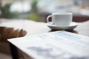 A newspaper with the word "BUSINESS" in blue at the top rests on a table. In the background, there's a blurred white coffee cup on a saucer. The setting appears to be a calm, casual café or home environment.