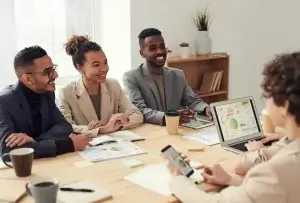 Four people are seated at a table in a meeting room. Three are viewing charts on a laptop, while one checks a smartphone. There are papers, mugs, and a plant on the table, and a shelf with decorations in the background.