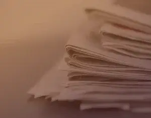 A stack of slightly crumpled newspapers or papers placed on a surface. The image has a warm, sepia-toned filter, giving it a vintage or nostalgic feel. The papers are layered unevenly, with the top sheets slightly curled.
