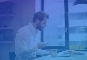 Man in a white shirt sitting at a desk, looking at a smartphone, with an open laptop in front of him. A coffee cup is on the table. The scene has a blue tint, and a large window is in the background.