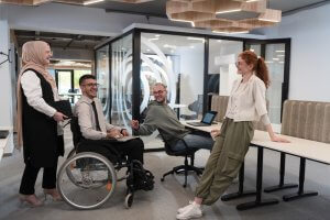 Four colleagues in a modern office, possibly working in the public sector, engage in a friendly discussion. One is in a wheelchair while others are seated or standing. They appear diverse in age and background, with a glass-walled meeting room visible behind them.