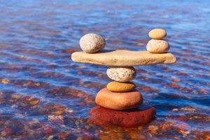 A balanced stack of smooth stones, with a large flat stone in the middle, stands in clear, rippling water. The stones range in color from red to beige, reflecting a serene truth and harmonious arrangement against the blue water.
