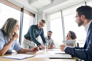 A diverse group of five people engages in a serious discussion in a modern office with large windows. One person takes the lead, standing and pointing at a document on the table, while others sit attentively, taking notes and engaging in conversation.