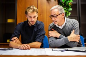 Two men sit at a table covered with papers, reviewing documents. The younger man with a beard wears a dark shirt, while the older man with glasses and gray hair sports a sweater. They appear focused on discussing public sector matters, deep in concentration and dialogue.