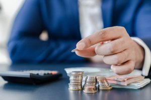 A person in a blue suit holds a coin over several stacked coins, symbolizing financial planning. In the background, a calculator and a pile of banknotes rest on a black surface, hinting at the complexities of government budgeting or economic calculation.