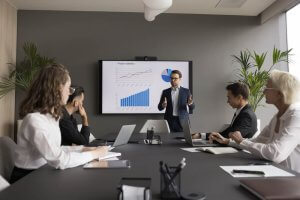 A man in a suit presents a business report on a large screen showing charts to his team of four colleagues seated at a conference table in a modern office. The room has plants, and laptops are visible on the table.